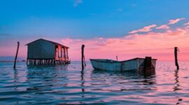 Deretan perahu kayu tradisional milik nelayan bersandar rapi di tepi permukiman panggung pesisir Indonesia saat senja menyapa.