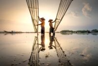 Deretan perahu kayu nelayan tradisional tertambat rapi di pinggir dermaga sebuah desa pesisir Indonesia saat matahari terbenam menyinari perairan pantai.