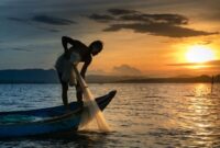 Seorang nelayan tradisional di atas perahu kayu bersiap melaut menyongsong matahari terbit, menggambarkan keharmonisan antara kehidupan masyarakat pesisir dan bentang alam bahari Nusantara.