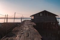 Deretan rumah panggung kayu warga pesisir yang berdiri di atas perairan jernih dengan latar belakangan pemandangan matahari terbenam. Arsitektur tradisional ini dirancang khusus untuk menyesuaikan diri dengan dinamika pasang surut air laut Nusantara.