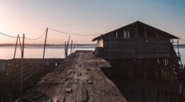 Deretan rumah panggung kayu warga pesisir yang berdiri di atas perairan jernih dengan latar belakangan pemandangan matahari terbenam. Arsitektur tradisional ini dirancang khusus untuk menyesuaikan diri dengan dinamika pasang surut air laut Nusantara.
